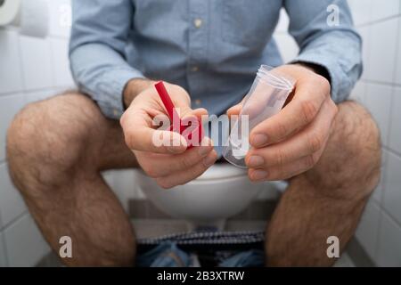 Stool Sample Container In Hands Of Man Sitting On Toilet Stock Photo