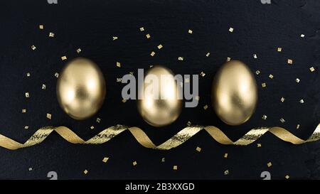 Golden egg with golden shine decorated confetti, ribbon for banner, greeting card, social media advertise. Gold eggs on black stone background. Flat l Stock Photo