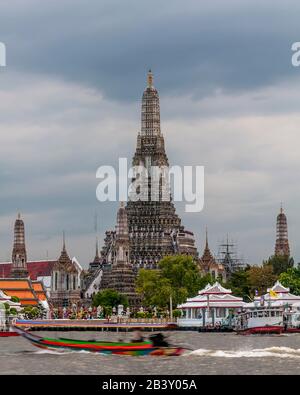 A colorful long tail boat sails on the Chao Phraya river in front of the Wat Arun temple, against cloudy sky, Bangkok, Thailand Stock Photo