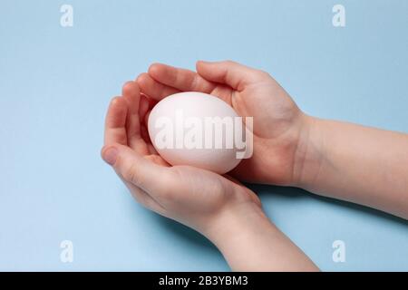 White chicken egg in the hands of a child on a blue background. Easter concept. Stock Photo