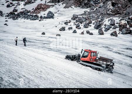 Close up of red snowcat (snow groomer) on glacier. Mount Elbrus, Caucasus, Russia.Two alpinist crossing before. Rocks in background. Stock Photo