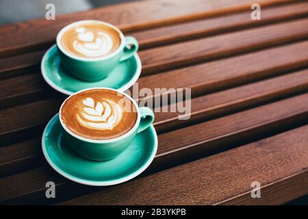 Two cups of cappuccino with latte art on wooden background. Beautiful foam, greenery ceramic cups, stylish toning, place for text. Stock Photo