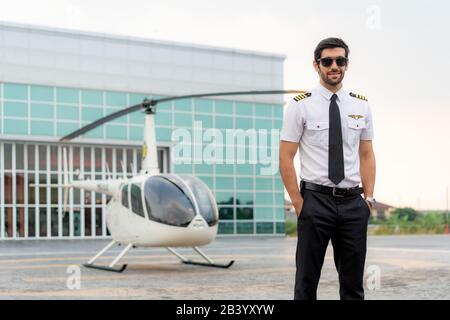 Portrait of handsome commercial pilot in captain white uniform standing and looking smart near small private helicopter on a landing point with privat Stock Photo