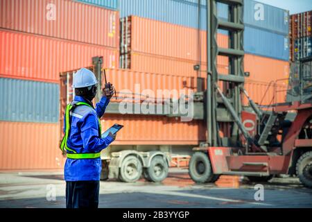 Foreman using and talking walkie talkie to control loading Containers box to truck at Container depot station for Logistic Import Export Background. B Stock Photo
