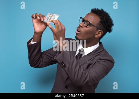 Serious young african american business man in suit and glasses standing, holding money checking it on fake. Studio shot on blue wall. Stock Photo