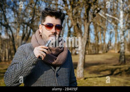Confident young bearded male in sunglasses vape electronic cigarette. A young handsome bearded hipster man vaping e-cigarette outdoors in the park Stock Photo