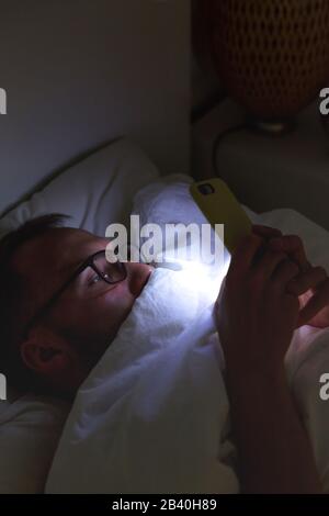 Addiction, nomophobia, insomnia. Close up of sleepy man in glasses using smartphone, lying on bed under the blanket at late night, can not sleep. Stock Photo