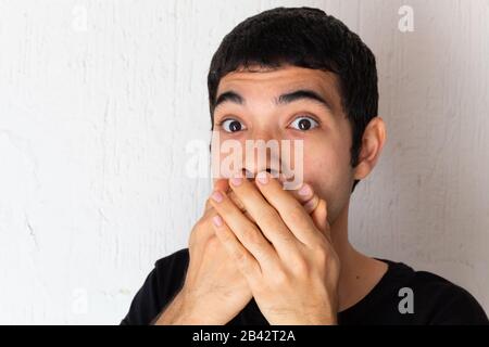 Young Hispanic man with black clothes surprised with his hands in his mouth Stock Photo