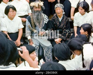 An elderly Ainu couple in their traditional dress pose with Japanese school girls visiting an Ainu village on the island of Hokkaido in northern Japan. The bearded man sits next to his wife with a faint tattoo resembling a mustache around her mouth. The painful tradition of facial tattooing during the childhood of Ainu girls was to indicate she was qualified for marriage.The practice has since been outlawed by the Japanese government. The old man and woman were two of only 300 pure-blooded Ainu (pronounced I-noo) still living when they posed for this historical photograph in 1962. Stock Photo
