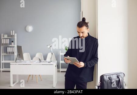 A serious guy is standing with a tablet in his hand while standing in the office at the workplace. Stock Photo