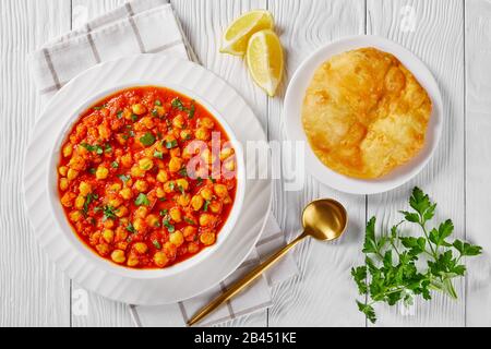 Chana masala or chickpea curry with spices, tomato sauce, served on a white plate with indian fried bread bhatura, lemon wedges,  on a wooden backgrou Stock Photo