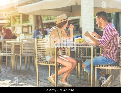 Happy couple eating piadina sandwich and toasting beer in bar kiosk restaurant. Young people having fast meal sitting outdoor - Relationship concept - Stock Photo