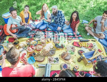 Semi flat view of friends making picnic in nature park sitting on grass- Young people having fun playing music, eating and drinking at bbq party - Mai Stock Photo