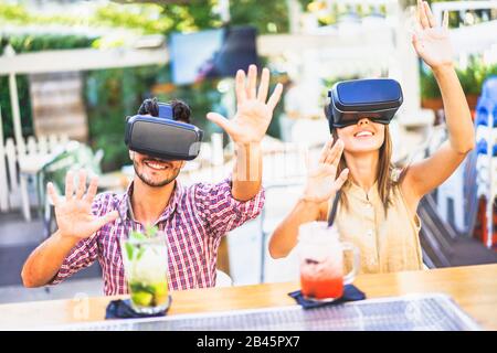 Amazed friends looking in VR goggles and gesturing with hands in cocktail bar restaurant - Young people having fun with new phone trends - Technology Stock Photo