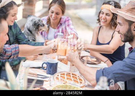 Happy friends cheering at breakfast brunch meal in nature outdoor - Young people having fun together eating fruits and drinking smoothies - Summer con Stock Photo