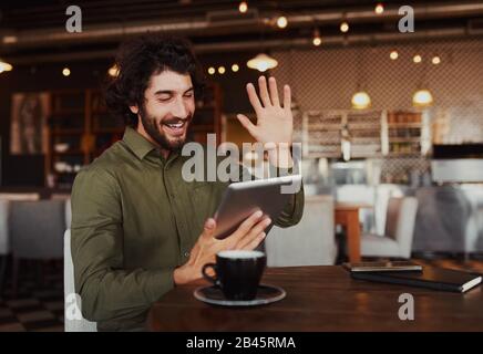 Casual bearded man waving his hand while talking through video-chat in smartphone while relaxing in cafe Stock Photo