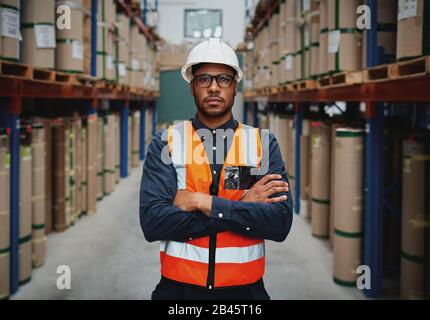 Confident young manager with arms crossed standing wearing orange vest and white hardhat in manufacturing unit looking at camera Stock Photo