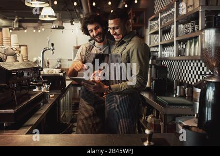 Smiling handsome waiter holding tablet with coworker while pointing on it in coffee shop sharing recipe Stock Photo