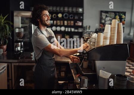 Young cheerful barista wearing black apron while preparing coffee at an automatic machine in a modern coffee shop Stock Photo