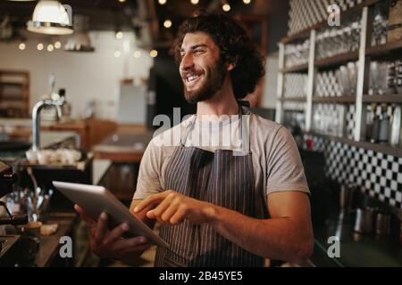 Portrait of successful young caucasian cafe owner standing behind counter using digital tablet