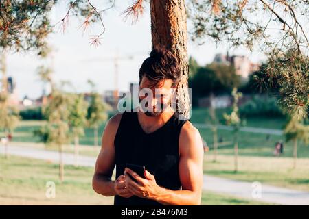 smiling attractive sporty man in tank top leaning against a tree looking at his mobile phone in workout break Stock Photo