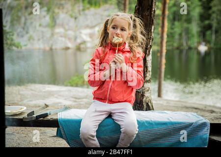 Man and his little daughter having barbecue in forest on rocky shore of lake, making a fire, grilling bread, vegetables and marshmallow. Happy Family Stock Photo