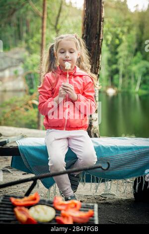 Man and his little daughter having barbecue in forest on rocky shore of lake, making a fire, grilling bread, vegetables and marshmallow. Happy Family Stock Photo