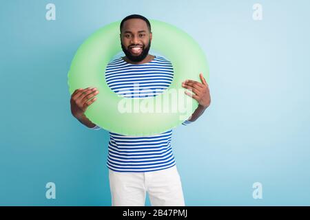 Photo of funny cheerful dark skin guy hold green life buoy around neck ready swim ocean sea traveler good mood sunny day wear striped sailor shirt Stock Photo