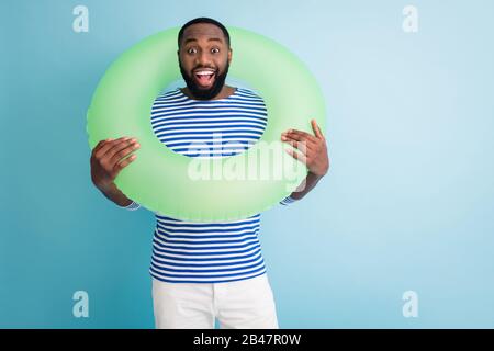 Photo of cheerful dark skin guy hold green life buoy around neck ready swim ocean sea traveler good mood sunny day wear striped sailor shirt isolated Stock Photo