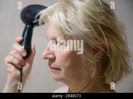 Aged woman dries hair with a hairdryer at home. Stock Photo