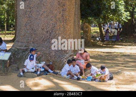 Kandy, Sri Lanka: 03/19/2019: Peradeniya Botanical Gardens school children and teacher seated under tree eating lunch. Stock Photo