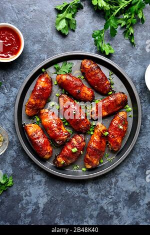 Bacon wrapped grilled chicken wings on plate over blue stone background. Tasty snack from chicken meat, bacon in sweet, sour, salty and spicy sauce. T Stock Photo