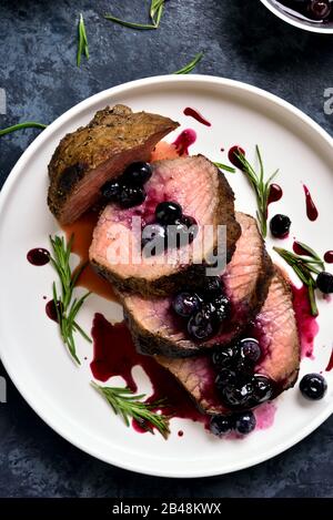 Close up of sliced grilled beef with blueberry sauce on white plate over blue stone background. Tasty medium rare roast beef with berry sauce. Top vie Stock Photo