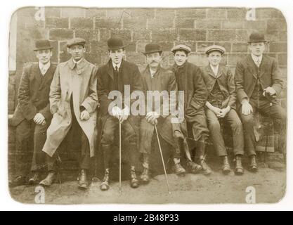 Early 1900's Edwardian cabinet card of group of seven young men and boys wearing flat caps and bowler hats, some holding walking sticks or canes, rounded collars, sitting together resting after a walk on a bench, U.K. Circa 1905 Stock Photo