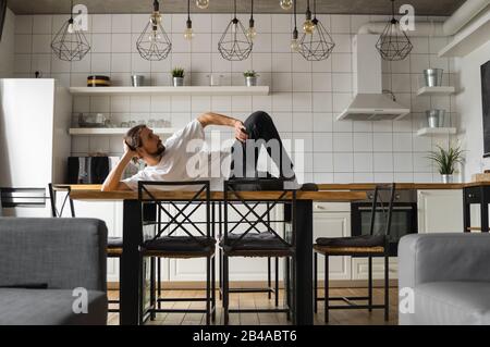 Young freelancer succeeded with project working from home and relaxing laying on the table at the kitchen. Successful bearded man Stock Photo