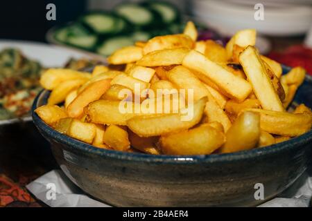 French fries served on a plate on table with blurred cucumbers on background, close-up. Stock Photo