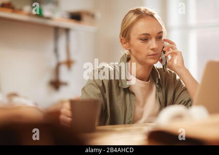 Attractive young woman in casual outfit sitting at table with cup of coffee and laptop on it talking on phone Stock Photo