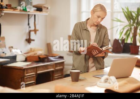 Horizontal shot of young female artisan writing down order details in her handmade notebook with leather cover Stock Photo
