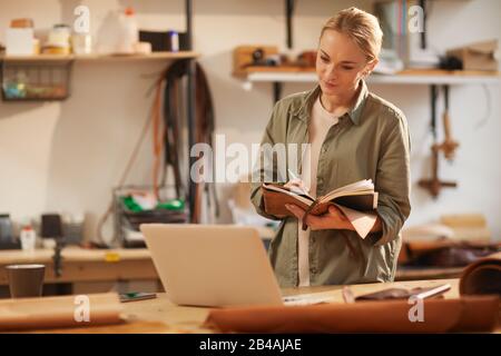 Horizontal shot of young female artisan looking at laptop screen writing something in her handmade notebook with leather cover Stock Photo