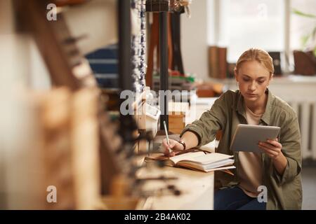 Horizontal shot of young blond woman sitting at table in craft workshop looking at tablet PC screen and writing down something in notebook Stock Photo