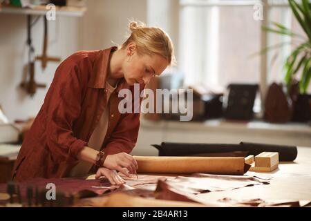 Horizontal side view medium shot of young Caucasian female artisan cutting out leather for new handiwork Stock Photo