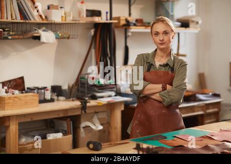 Horizontal shot of confident young woman wearing apron standing at work table with with arms crossed looking at camera Stock Photo