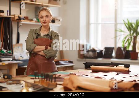Horizontal portrait of confident Caucasian woman wearing apron standing with arms crossed in modern leather craft workshop looking at camera, copy spa Stock Photo