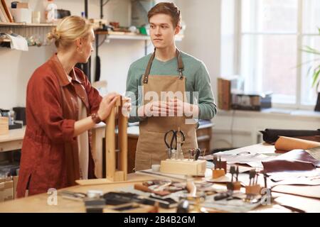 Caucasian female artisan demonstrating her apprentice how to use leather craft wooden clip, horizontal shot Stock Photo