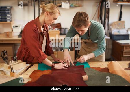 Young man creating stitching holes in leather material while woman stabilizing it on work surface Stock Photo