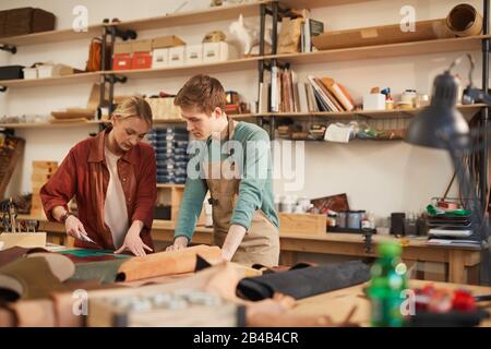 Attentive young man and concentrated woman creating new leather handicraft together in modern workshop Stock Photo