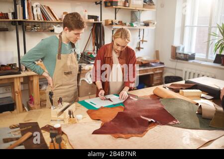 Horizontal shot of young adult female artisan working with leather detail, her colleague looking at her Stock Photo