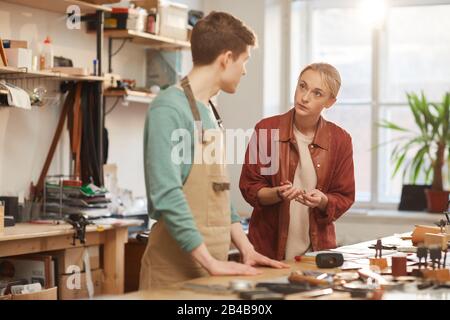Horizontal shot of young man and woman working together in leather craft workshop standing at table discussing something Stock Photo