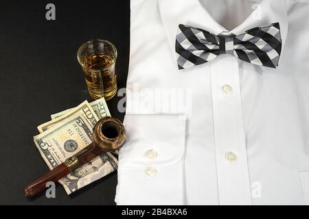 white dress shirt and black and white checkered bow tie with shot of whiskey and smoking pipe on American money Stock Photo