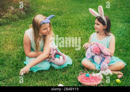 Cute little children wearing bunny ears on Easter day sitting on grass in garden and playing with painted eggs and Easter bunny toy. Stock Photo
